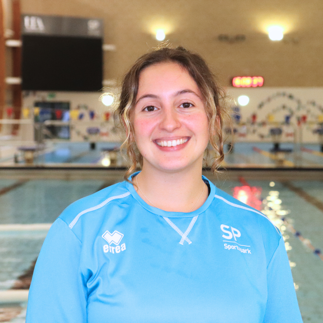 A smiling young woman with light brown curly hair in a blue athletic top stands in front of an indoor swimming pool.