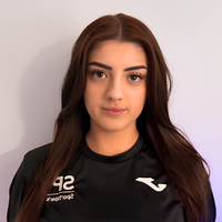 A young woman with long brown hair and brown eyes looks at the camera. She is wearing a black athletic jersey with SP and Joma logos.