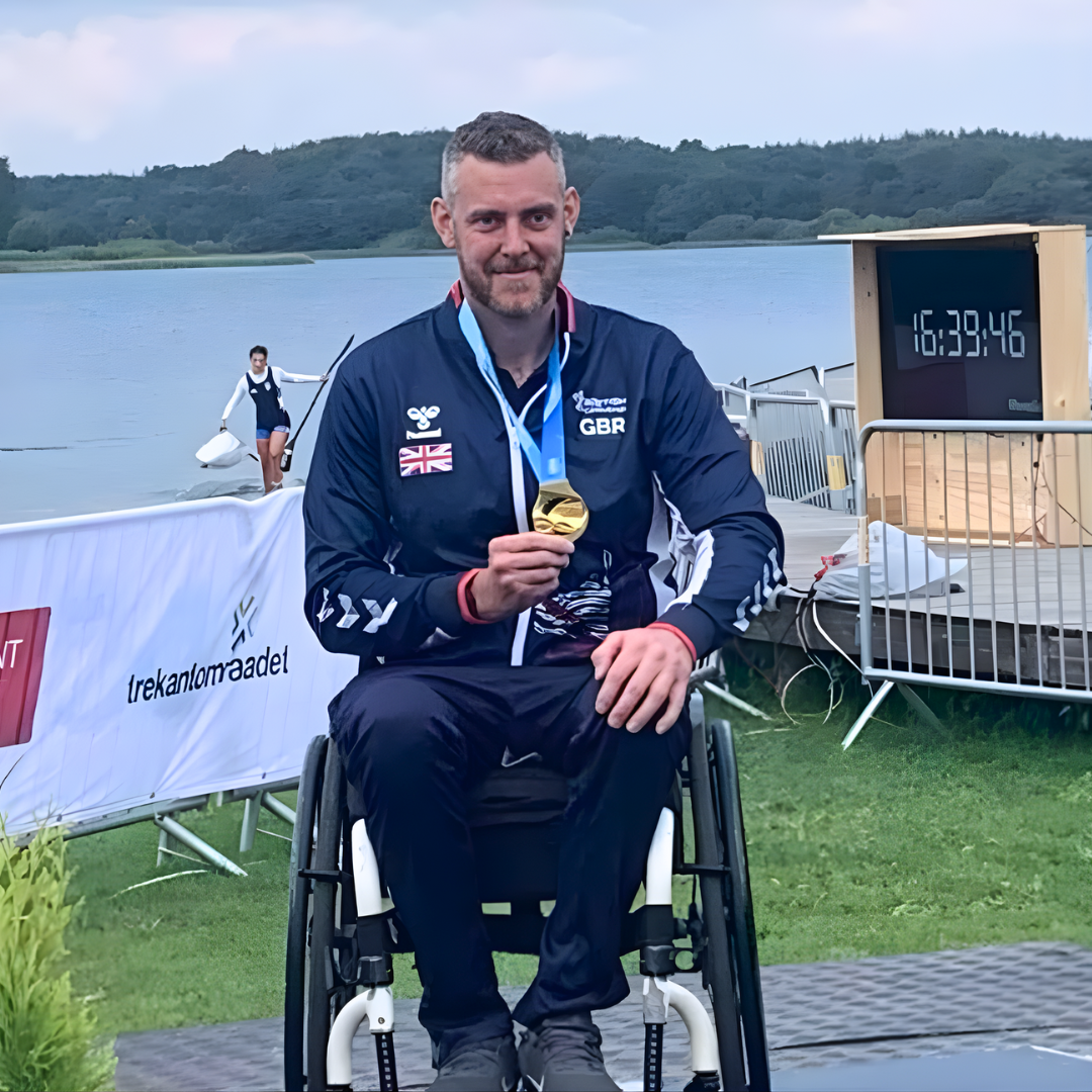 Shaun in wheelchair holding a gold medal in front of lake.