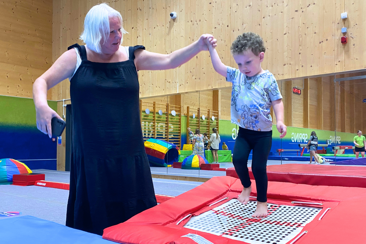 Older lady holding the hand of a young boy as he jumps on a small indoor trampoline in gymnastics centre.