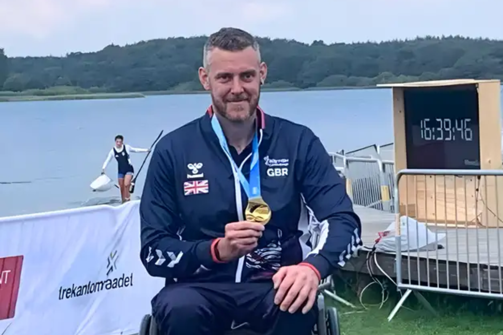 Shaun Cook with medal smiling at camera in front of lake.