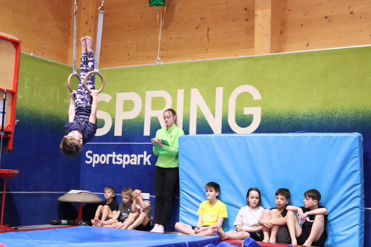 Boy hangs upside on gymnastics rings while coach scores him from the side with a clipboard.