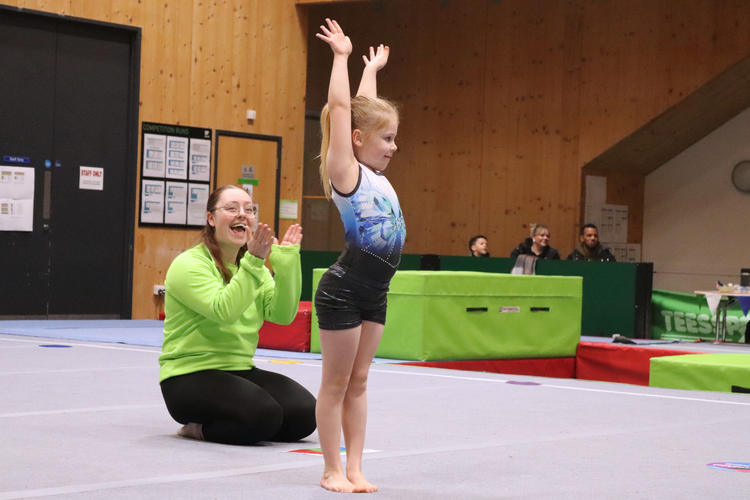 Small girl finishes gymnastics routine with arms in the air. her coaches kneels off to the side smiling and clapping,