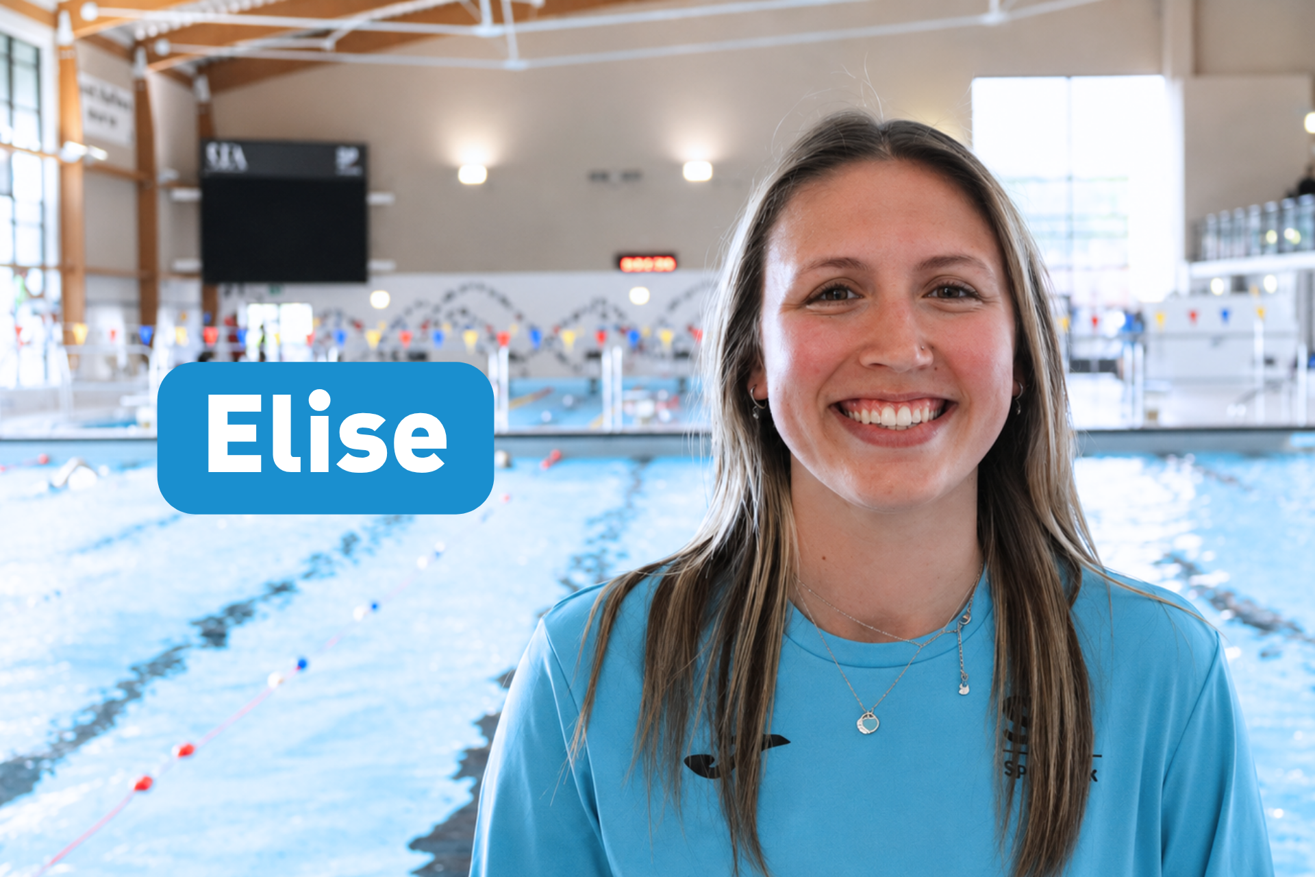 A smiling woman named Elise, wearing a blue shirt, poses for a photo in front of an indoor swimming pool.