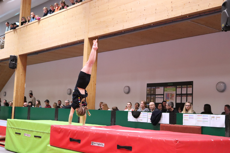 Girl doing handstand on safety mat in gymnastics centre while audience watches.