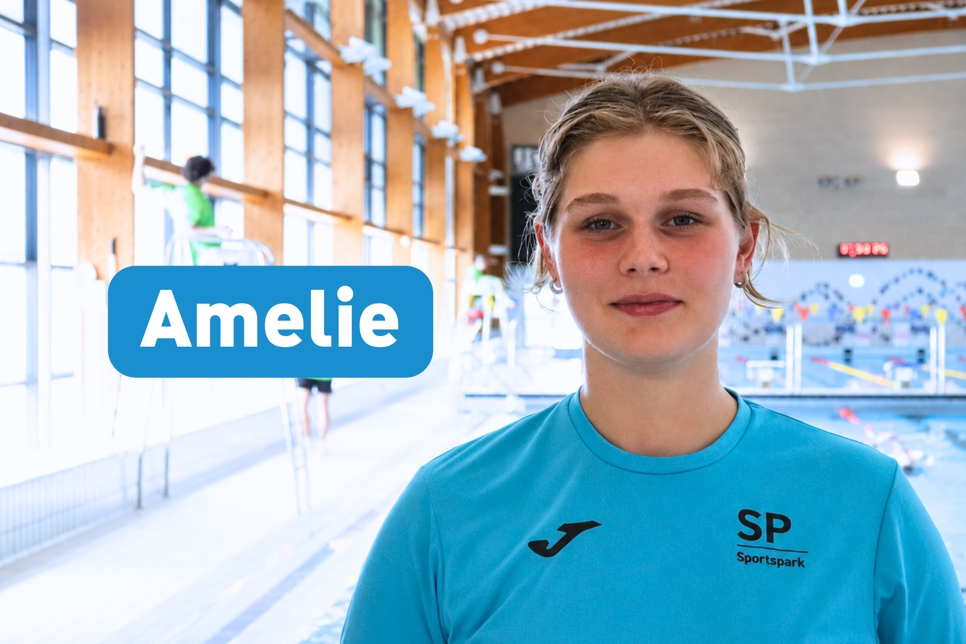 A young woman named Amelie in a blue Sportspark t-shirt looks at the camera, standing in a brightly lit indoor swimming pool hall.