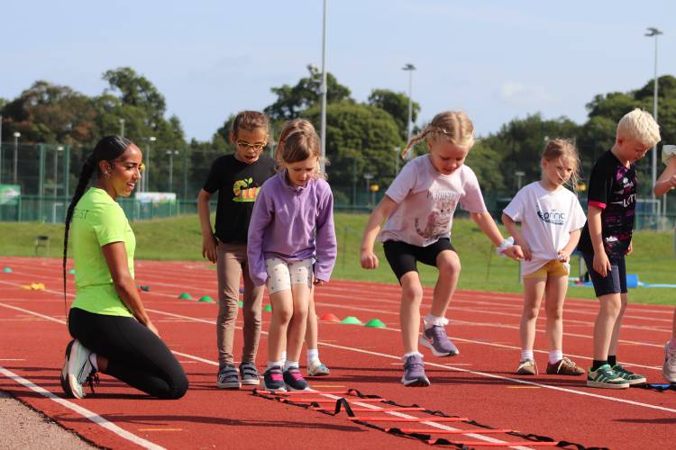 Sportspark instructor kneeling on athletics track while group of children run single file through athletics ladder.