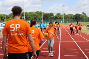 A team of one man and two girls in orange t-shirt competing in an egg and spoon race on athletics track.