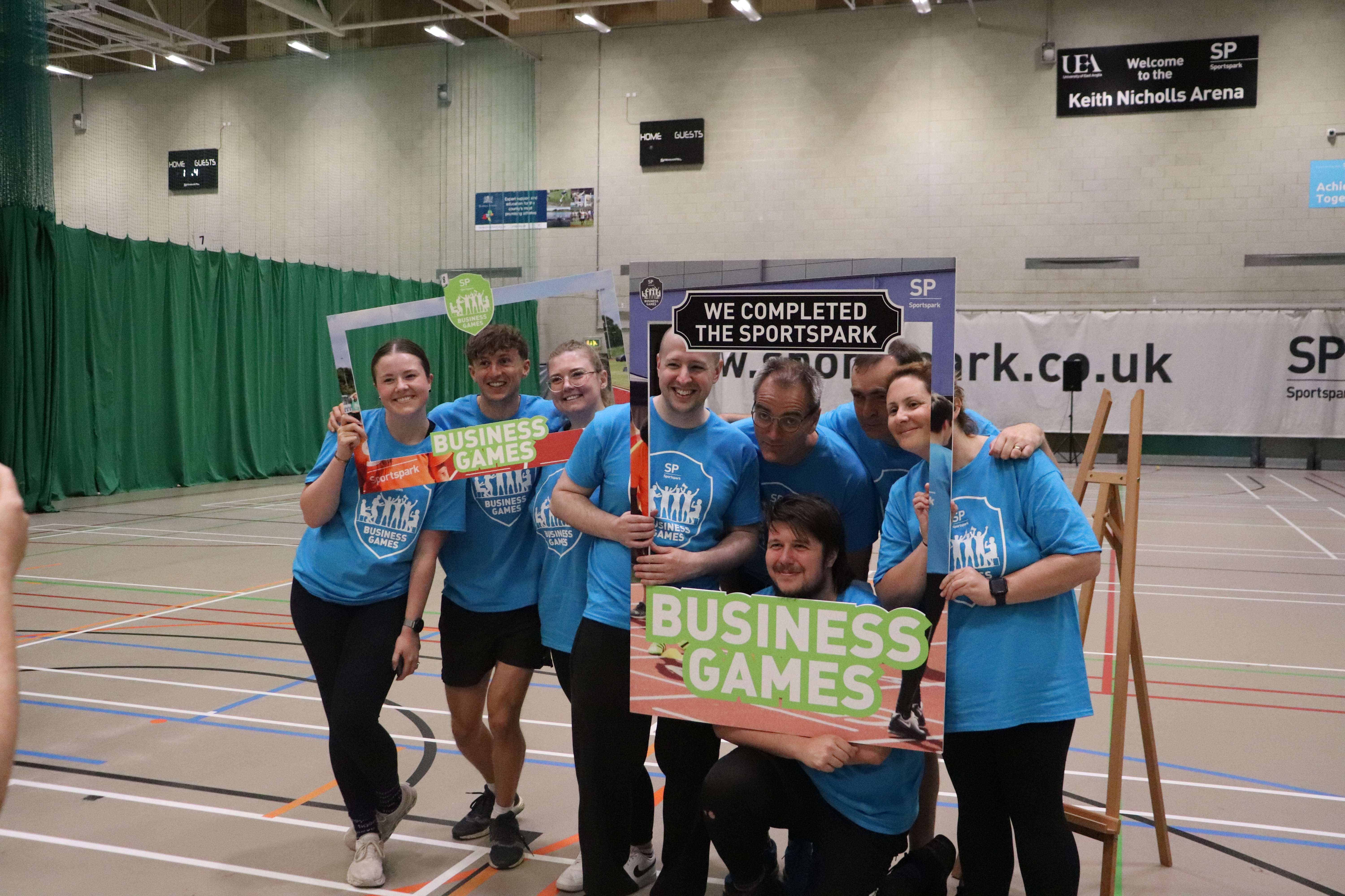 Group of people in blue t-shirts in sports hall taking a group photo with a selfie frame reading 'business games'